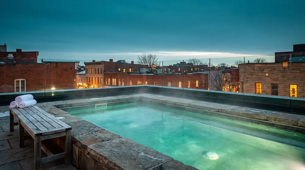 Generated illustration: steam rising from a turquoise rooftop mineral pool at dusk above the brick rooftops of a small prairie downtown